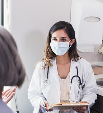Nurse Practitioner Pic An advanced practice provider wearing a white coat and a surgical mask talking to a patient inside of a medical office.